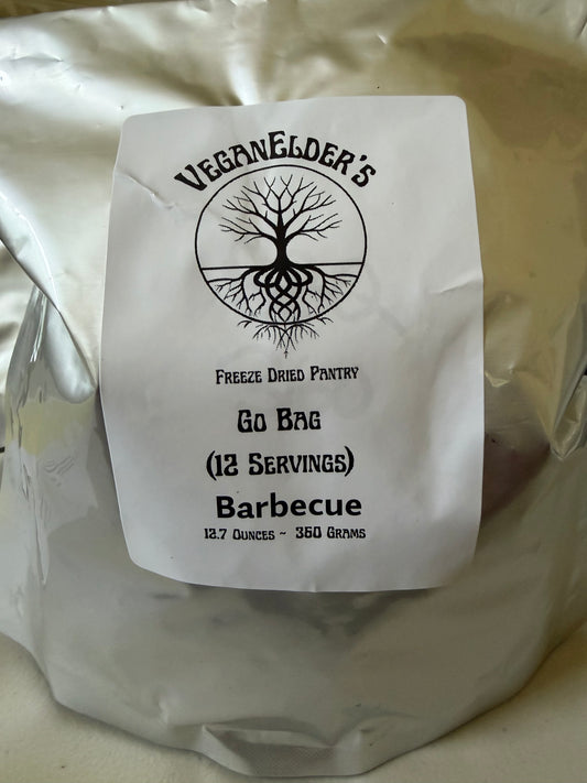 A silver foil bag of 12 servings of VeganeLder's Freeze Dried Pantry Barbecue Go Bag, featuring a white paper label with a black tree logo and text.