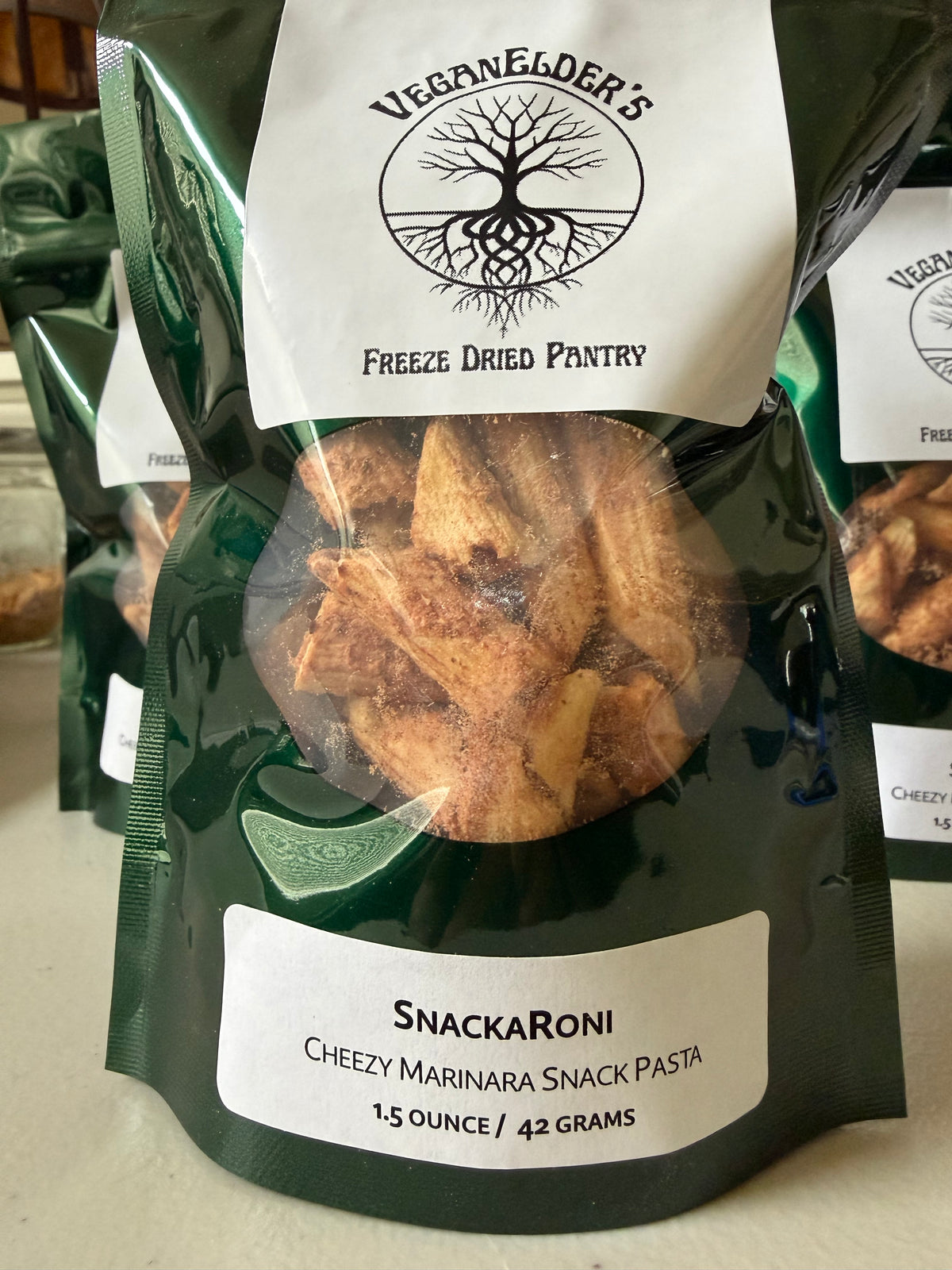 A bag of Veganelder's Freeze Dried Pantry SnackRoni Cheezy Marinara Snack Pasta, featuring a dark green and white packaging design with a clear window showing the freeze-dried pasta inside.