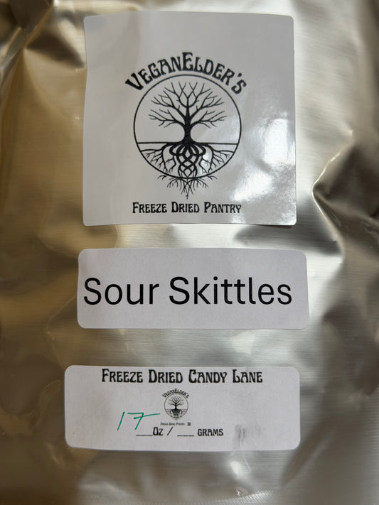 A close-up of a metallic silver bag of freeze dried Sour Skittles Candy with three white labels. The top label shows the logo for VeganElder's Freeze Dried Pantry, the middle label reads 'Sour Skittles', and the bottom label shows the VeganElder's logo and weight information.