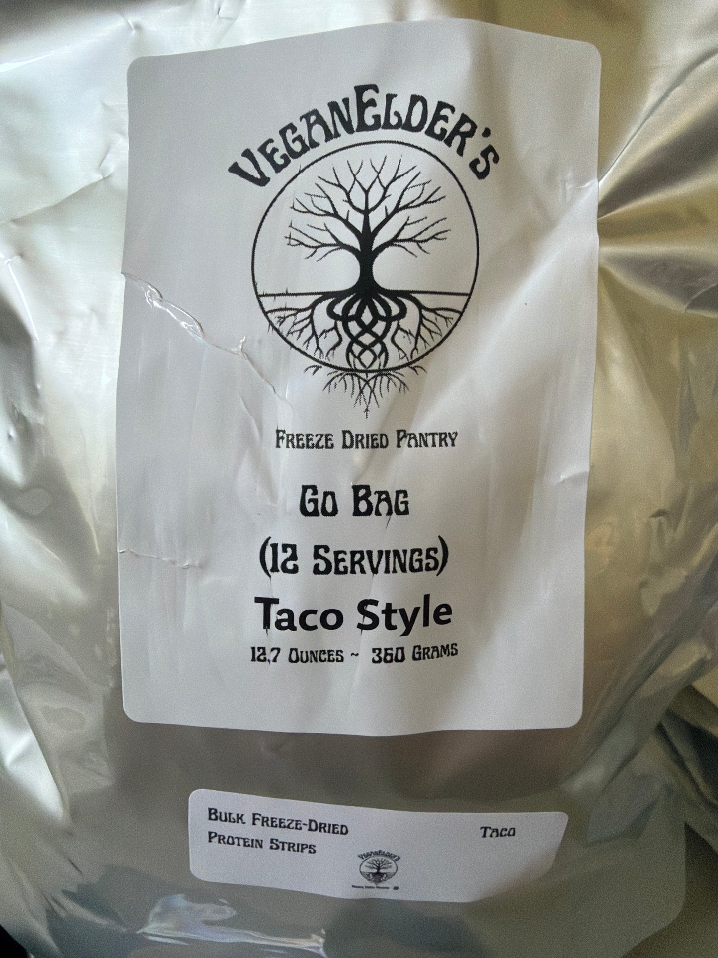 A silver bulk bag of Veganelder's Freeze Dried Pantry 12 servings Go Bag Taco Style Protein strips, featuring a white label with a black tree logo and text, and a smaller label for bulk freeze-dried protein strips.