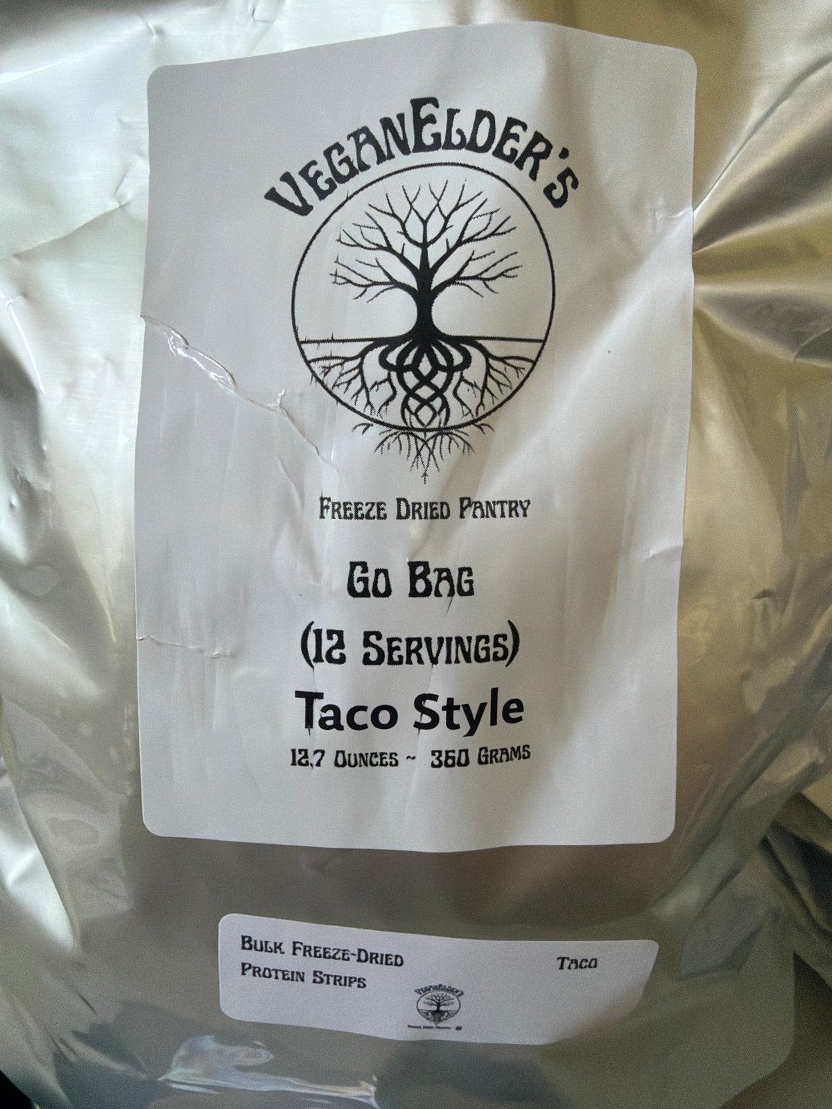 A silver bulk bag of Veganelder's Freeze Dried Pantry 12 servings Go Bag Taco Style Protein strips, featuring a white label with a black tree logo and text, and a smaller label for bulk freeze-dried protein strips.