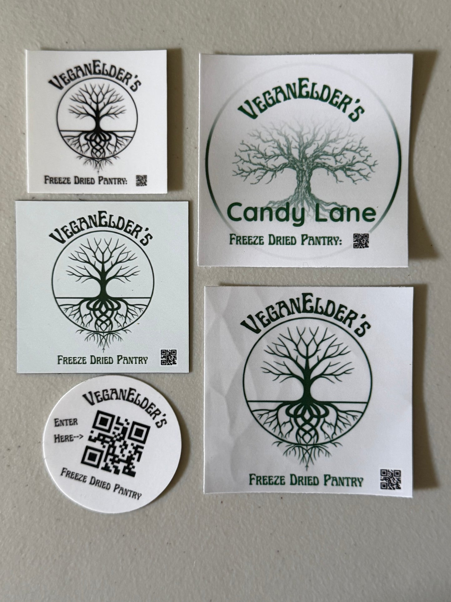 Five square and round white stickers with green and black Veganelder's branding and QR codes, arranged on a beige wall.