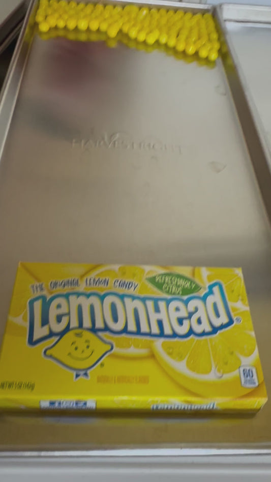 video of yellow candy balls rolling across a freeze drying tray with a lemondhead candy box in the foreground